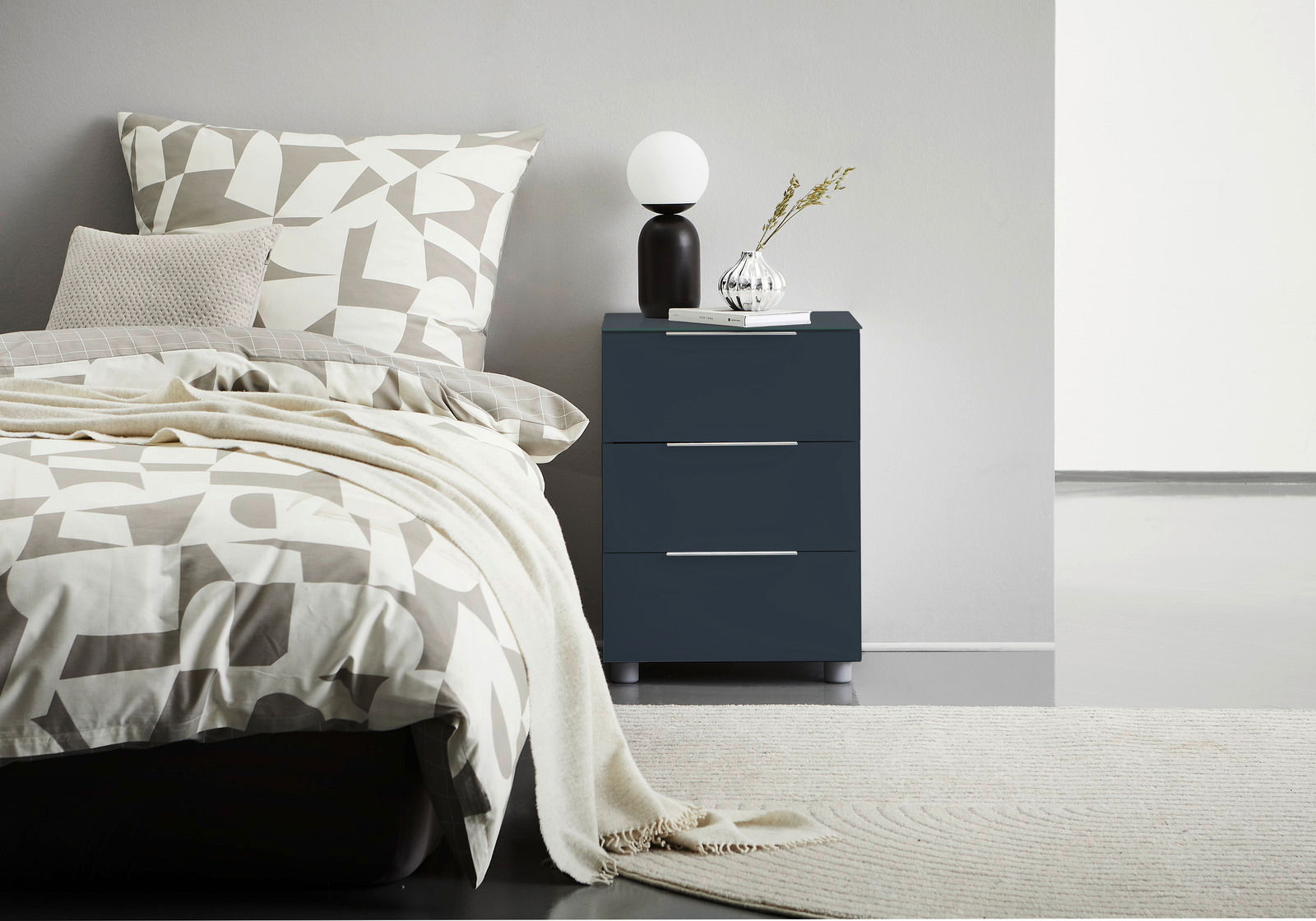 Dunkelblaue Nachtkonsole mit drei Schubladen, darauf eine Lampe und eine Vase mit Zweigen, seitliche Perspektive.