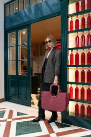 Frau mit Sonnenbrille und grauem Blazer steht in einer Türöffnung, hält eine große, rote Tasche in der Hand. Die Perspektive zeigt die Tasche von vorne.