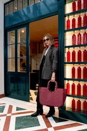 Frau mit Sonnenbrille und grauem Blazer steht in einer Türöffnung, hält eine große, rote Tasche in der Hand. Die Perspektive zeigt die Tasche von vorne.