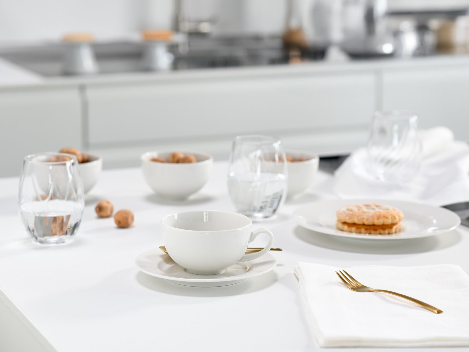 Weiße Kaffeetasse mit Untertasse auf einem hellen, modern gedeckten Tisch mit Wassergläsern, Snackschalen und goldfarbenem Besteck; Perspektive: leicht seitliche Tischhöhe mit Fokus auf die Tasse im Vordergrund.