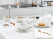 Weiße Kaffeetasse mit Untertasse auf einem hellen, modern gedeckten Tisch mit Wassergläsern, Snackschalen und goldfarbenem Besteck; Perspektive: leicht seitliche Tischhöhe mit Fokus auf die Tasse im Vordergrund.