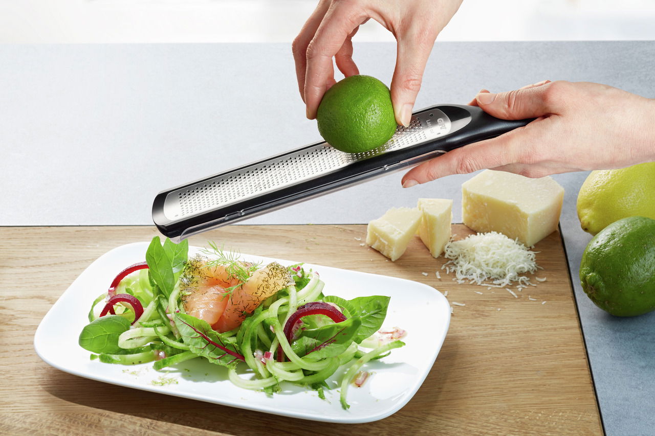 Feinreibe RAFINO in der Hand, die eine Limette über einem frischen Salat reibt, mit Käse und Limetten im Hintergrund. Perspektive von oben.