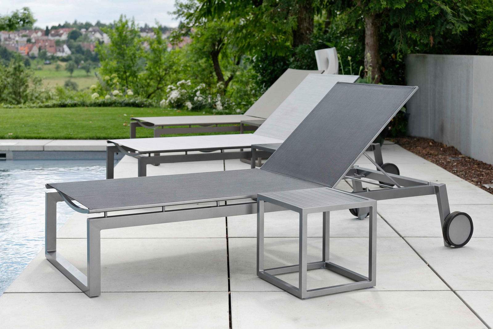 Moderne Rollenliege aus Metall mit verstellbarer Rückenlehne, seitliche Perspektive, auf einer Terrasse neben einem Pool mit grüner Landschaft im Hintergrund.