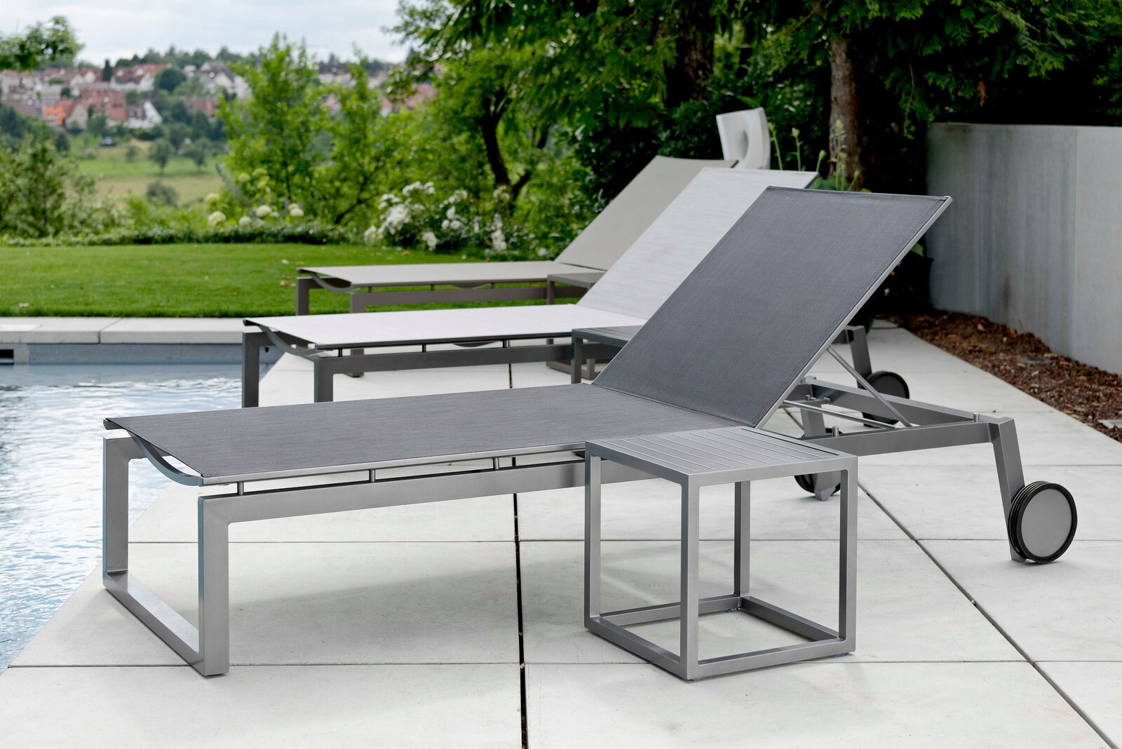 Moderne Rollenliege aus Metall mit verstellbarer Rückenlehne, seitliche Perspektive, auf einer Terrasse neben einem Pool mit grüner Landschaft im Hintergrund.