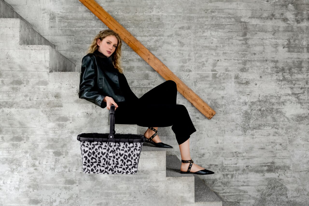 Reisenthel Carrybag Frame Leo Nero Frau sitzt auf einer Treppe und hält einen Einkaufskorb mit Leopardenmuster in der Hand, seitliche Perspektive.
