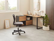 Moderner Winkelschreibtisch in einem hellen Raum, mit einem schwarzen Metallrahmen und Holzoberfläche, seitlich von einem Fenster beleuchtet. Ein ergonomischer Bürostuhl steht davor, und eine Pflanze in einem weißen Korb ergänzt das minimalistische Design