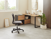 Moderner Winkelschreibtisch in einem hellen Raum, mit einem schwarzen Metallrahmen und Holzoberfläche, seitlich von einem Fenster beleuchtet. Ein ergonomischer Bürostuhl steht davor, und eine Pflanze in einem weißen Korb ergänzt das minimalistische Design