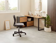 Moderner Winkelschreibtisch in einem hellen Raum, mit einem schwarzen Metallrahmen und Holzoberfläche, seitlich von einem Fenster beleuchtet. Ein ergonomischer Bürostuhl steht davor, und eine Pflanze in einem weißen Korb ergänzt das minimalistische Design