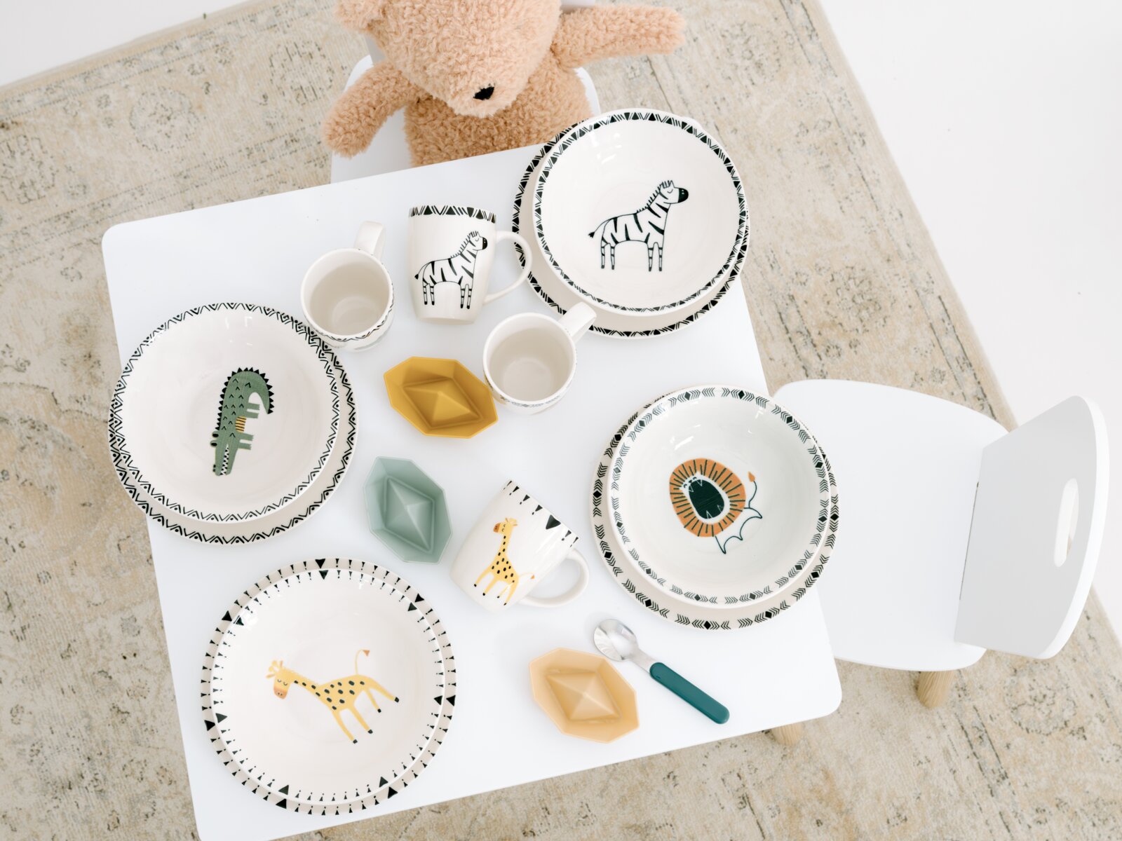 Kinder-Geschirrset mit Tellern, Schalen und Tassen in Weiß, verziert mit Zebra, Krokodil, Löwe und Giraffe, aus der Vogelperspektive auf einem weißen Kindertisch arrangiert