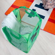 Grüner, faltbarer Aufbewahrungs- und Wäschekorb mit Netzseiten und Tragegriffen im Pixel-Design, von oben schräg fotografiert, mit hellgrünem Stoff darin auf hellem Boden neben einer orange-weißen Matte.