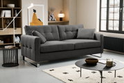 Modernes graues Stoffsofa mit Steppung, breiten Armlehnen und verchromten Füßen im Wohnzimmer, perspektivisch in leichter Frontansicht von links.