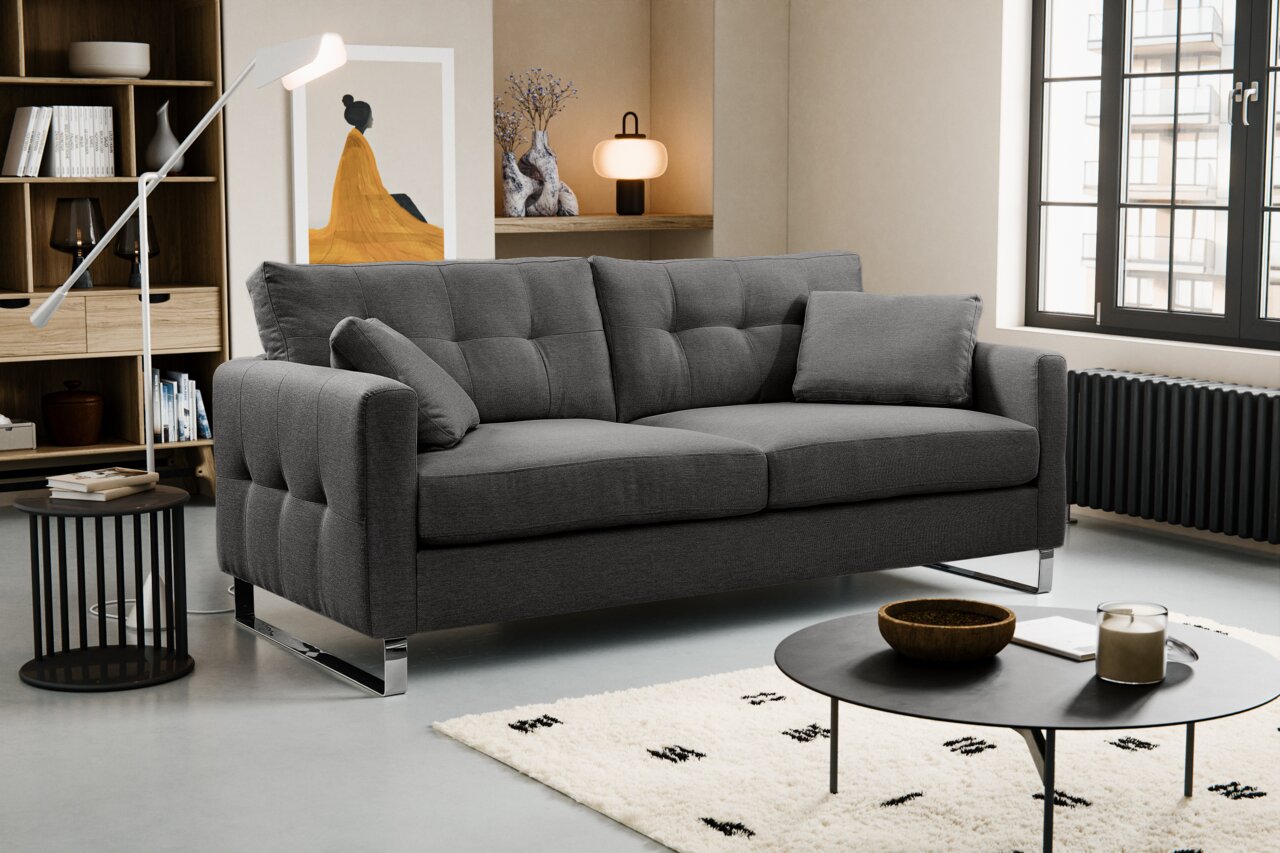 Modernes graues Stoffsofa mit Steppung, breiten Armlehnen und verchromten Füßen im Wohnzimmer, perspektivisch in leichter Frontansicht von links.