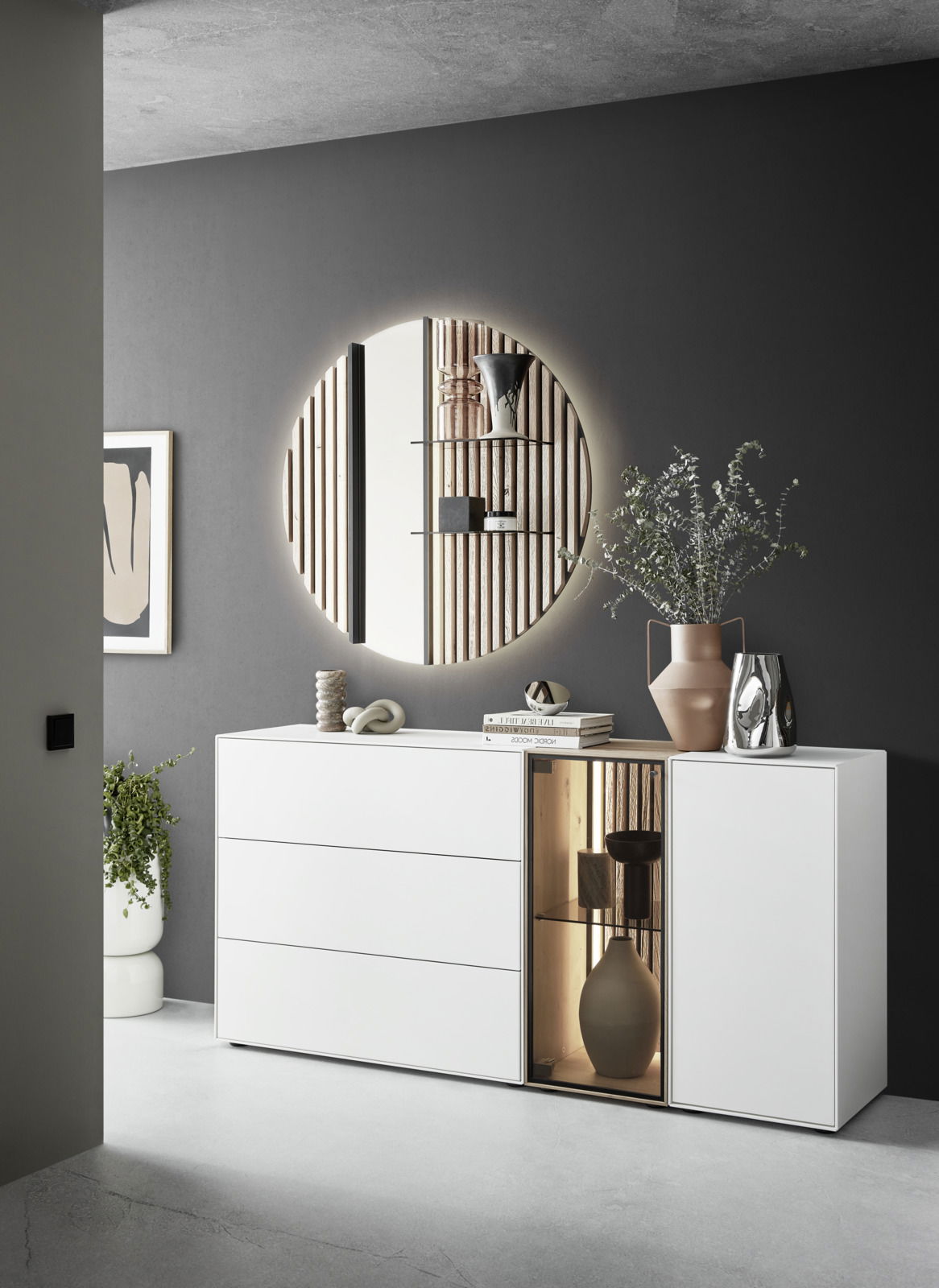 Modernes Sideboard mit Glasvitrine rechts, dekoriert mit Büchern und Vasen, in einem stilvollen Raum mit rundem Spiegel an der Wand. Perspektive von vorne links.