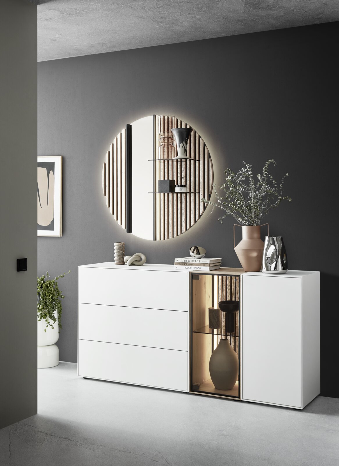 Modernes Sideboard mit Glasvitrine rechts, dekoriert mit Büchern und Vasen, in einem stilvollen Raum mit rundem Spiegel an der Wand. Perspektive von vorne links.