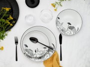 Draufsicht auf ein elegantes weißes Porzellan-Geschirrset mit schwarzem Blumenmuster, bestehend aus tiefem Teller/Schale und flachem Teller, dazu schwarzes Besteck, Glas und gelbes Tuch auf hellem Tisch
