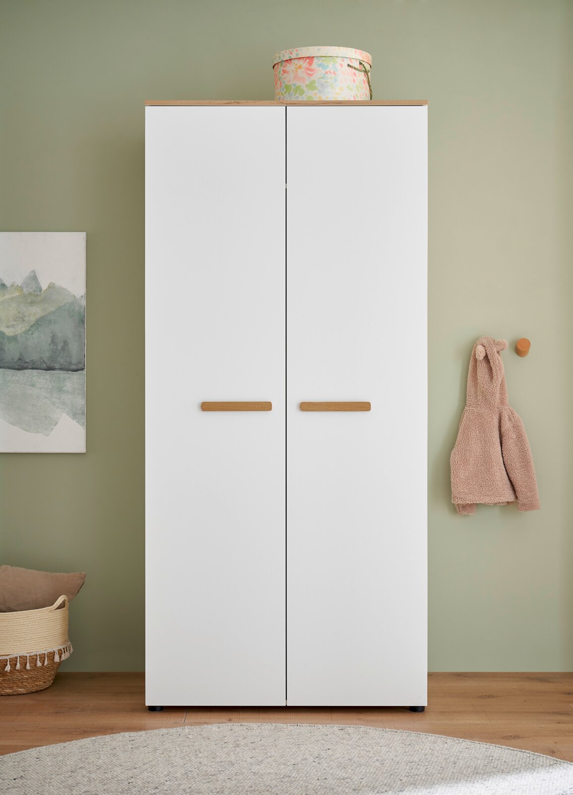 Weißer zweitüriger Kleiderschrank mit Holzgriffen und Holzdeckplatte, frontal von vorn in einem Raum mit grüner Wand aufgenommen.