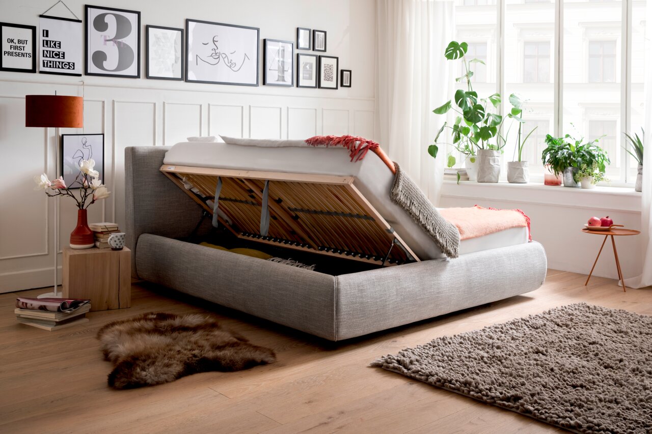 Seitliche Ansicht eines grauen Polsterbetts mit hochgeklappter Matratze, das Stauraum darunter zeigt. Das Bett steht in einem hellen, modern eingerichteten Raum mit Pflanzen und Wanddekorationen.
