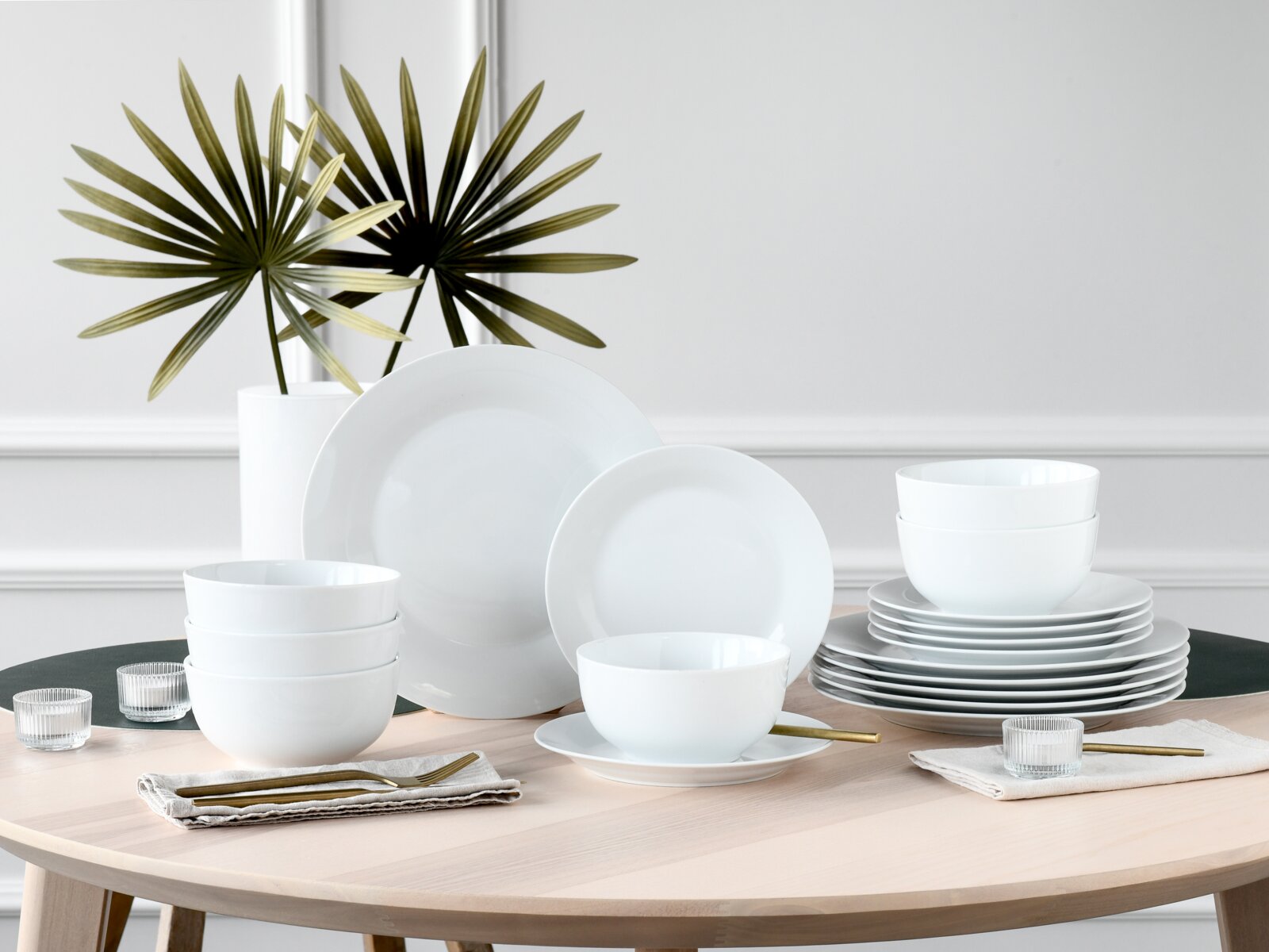 Weißes, minimalistisches Porzellan-Geschirrset mit Tellern und Schalen auf hellem Holztisch, frontale leicht erhöhte Perspektive im Wohnraum-Setting