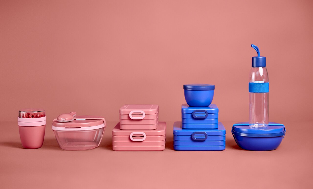Verschillende lunchboxen en bakjes in roze en blauw, vooraanzicht op een roze achtergrond.