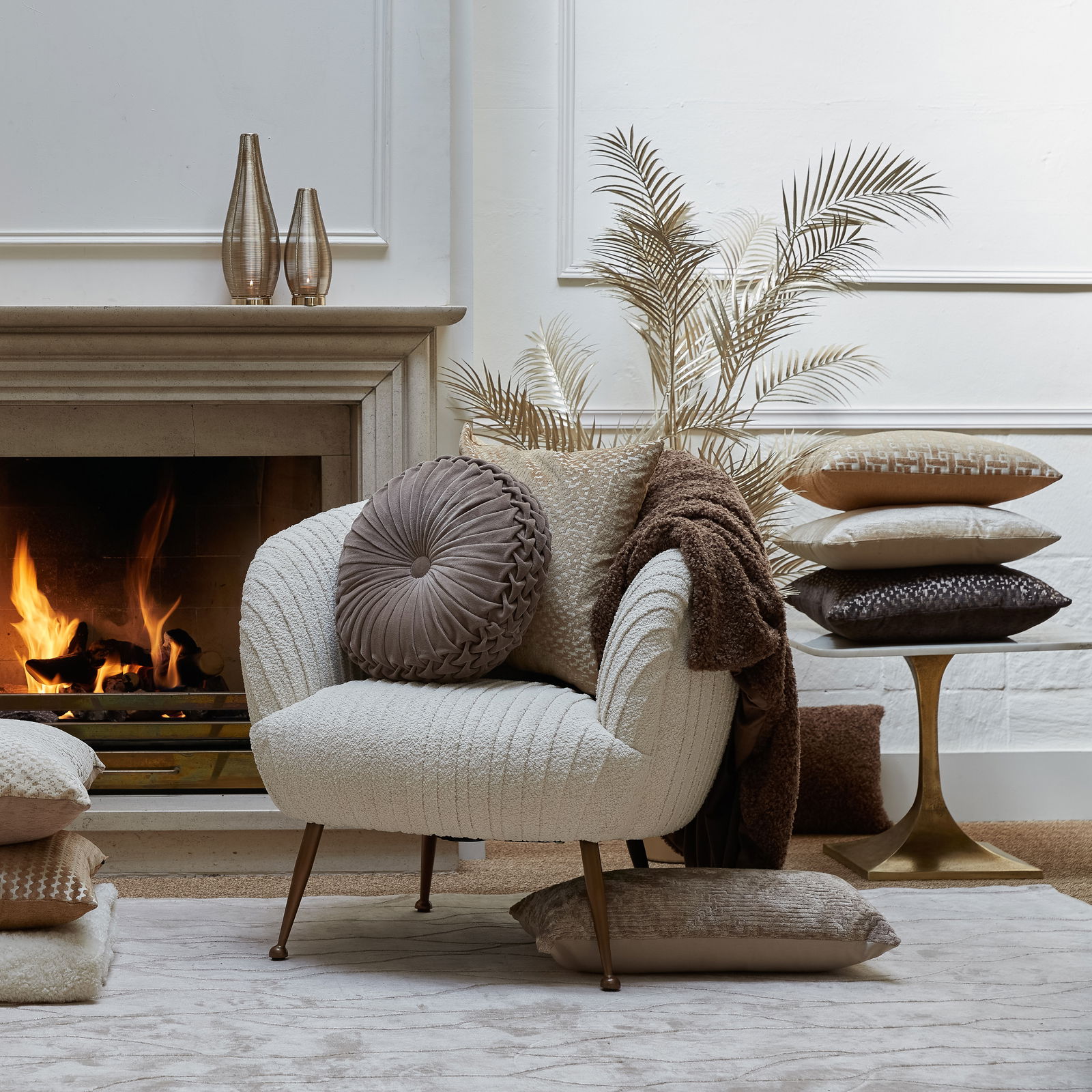 Gemütliche Wohnzimmer-Szene mit einem beigen Sessel, dekoriert mit verschiedenen Kissen in Erdtönen, vor einem Kamin. Perspektive von vorne.