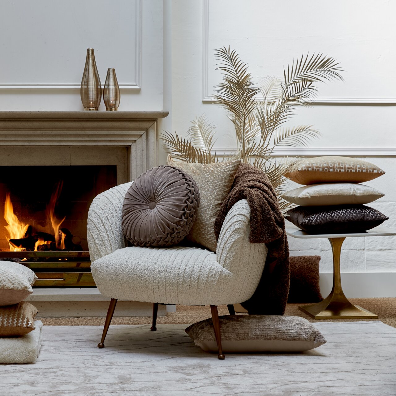 Gemütliche Wohnzimmer-Szene mit einem beigen Sessel, dekoriert mit verschiedenen Kissen in Erdtönen, vor einem Kamin. Perspektive von vorne.