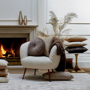 Gemütliche Wohnzimmer-Szene mit einem beigen Sessel, dekoriert mit verschiedenen Kissen in Erdtönen, vor einem Kamin. Perspektive von vorne.