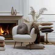 Gemütliche Wohnzimmer-Szene mit einem beigen Sessel, dekoriert mit verschiedenen Kissen in Erdtönen, vor einem Kamin. Perspektive von vorne.