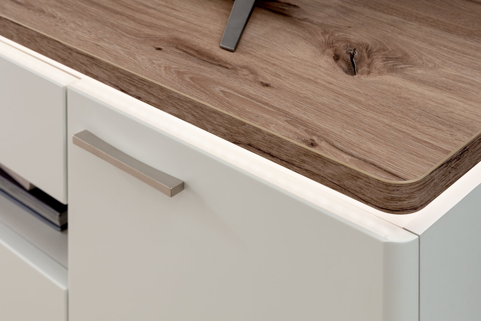 Detailfoto vanuit een schuin zijaanzicht: wit keukenfront met metalen handgreep en werkblad in houtlook met afgeronde rand