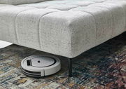 Eckgarnitur in hellem Stoffbezug mit einem Saugroboter, der darunter fährt, aus seitlicher Perspektive.