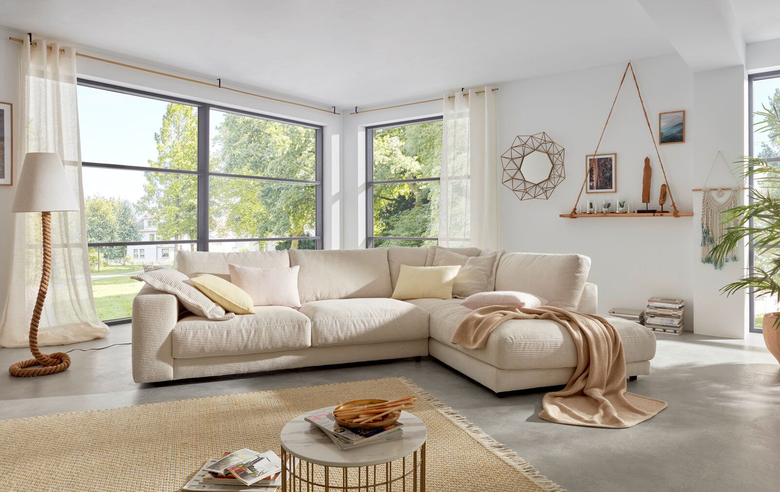 Gemütliche Rundecke in einem hellen Wohnzimmer mit großen Fenstern, dekoriert mit Kissen und Decke, aus der Perspektive von vorne rechts.