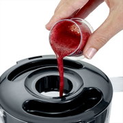 Close-up van bovenaf: hand giet rode smoothie/vruchtensap uit een maatbekertje in de opening van het zwarte deksel van een blender/keukenmachine.