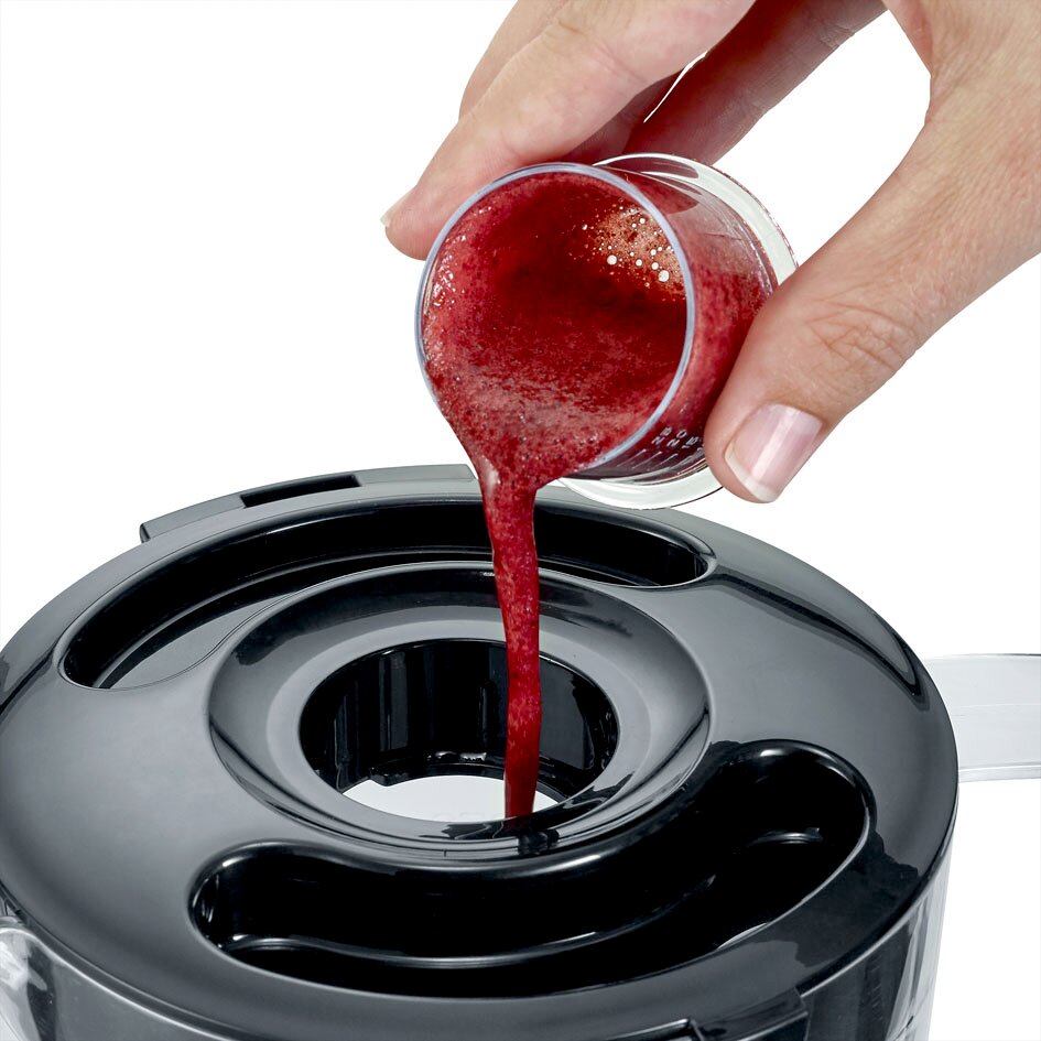 Close-up van bovenaf: hand giet rode smoothie/vruchtensap uit een maatbekertje in de opening van het zwarte deksel van een blender/keukenmachine.