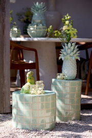 Zwei grüne, geflieste Hocker im Freien, einer mit einer Ananasvase mit Blumen und der andere mit einer grünen Papageienfigur und einem Glasobjekt. Perspektive von vorne.
