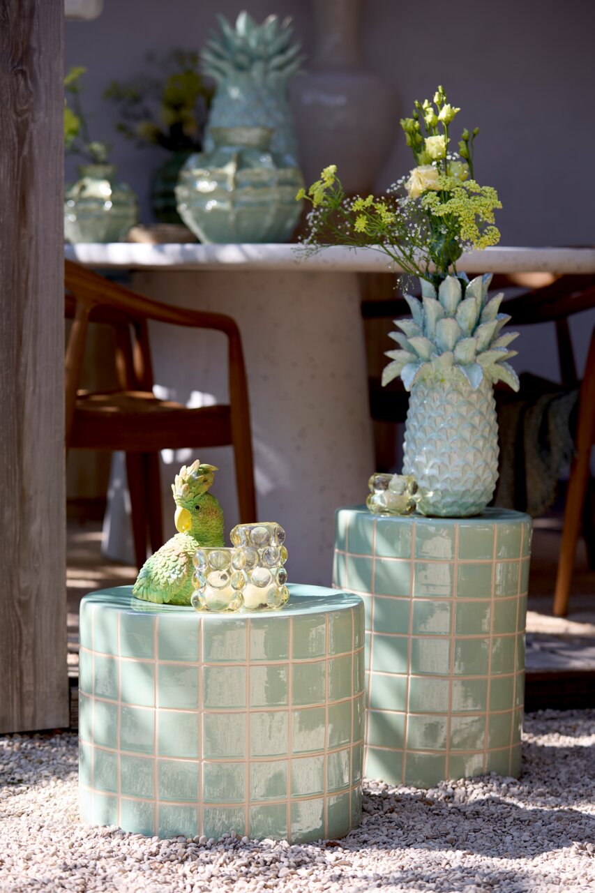 Zwei grüne, geflieste Hocker im Freien, einer mit einer Ananasvase mit Blumen und der andere mit einer grünen Papageienfigur und einem Glasobjekt. Perspektive von vorne.
