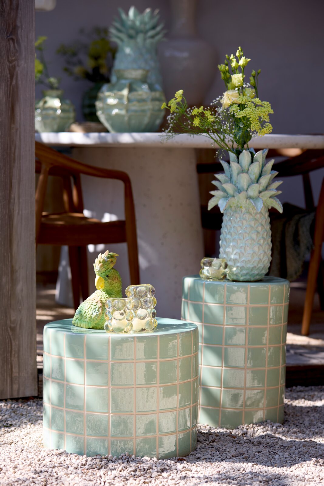 Zwei grüne, geflieste Hocker im Freien, einer mit einer Ananasvase mit Blumen und der andere mit einer grünen Papageienfigur und einem Glasobjekt. Perspektive von vorne.