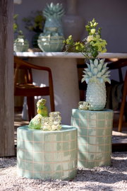 Zwei grüne, geflieste Hocker im Freien, einer mit einer Ananasvase mit Blumen und der andere mit einer grünen Papageienfigur und einem Glasobjekt. Perspektive von vorne.