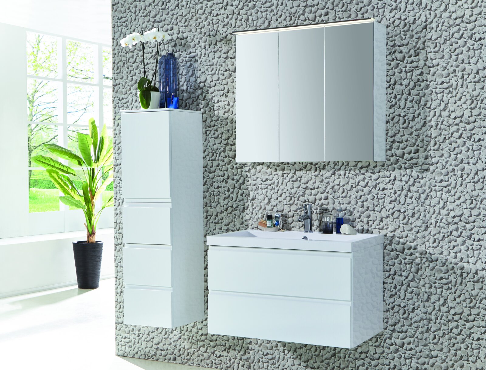 Modernes weißes Badezimmermöbel-Set mit Waschbecken-Unterschrank, seitlichem Hochschrank und dreitürigem Spiegelschrank an grauer Steinwand, Frontansicht in leichter Schrägperspektive von links