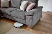Grijze hoekbank met kussens, gezien vanaf de zijkant, op een houten vloer met daaronder een robotstofzuiger.