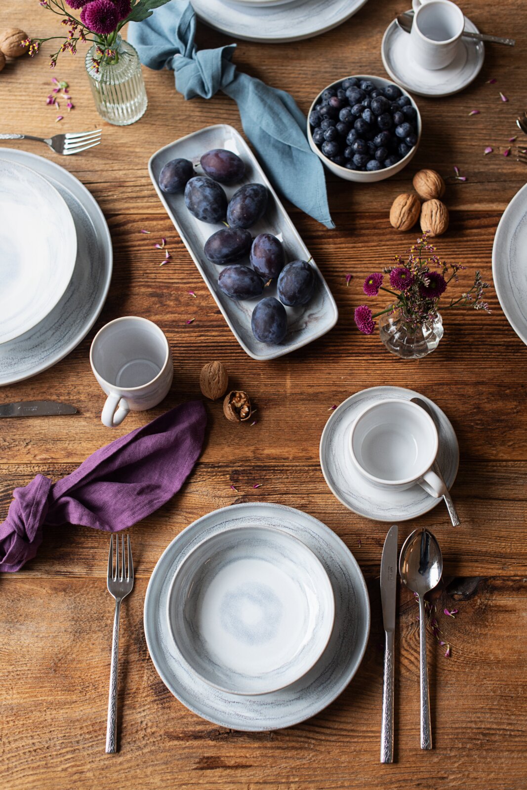 Draufsicht auf einen rustikal gedeckten Holztisch mit graublauem Keramikgeschirr, Tassen, Besteck, Stoffservietten sowie Schalen mit Blaubeeren und Pflaumen, dekoriert mit kleinen violetten Blumen und Walnüssen.