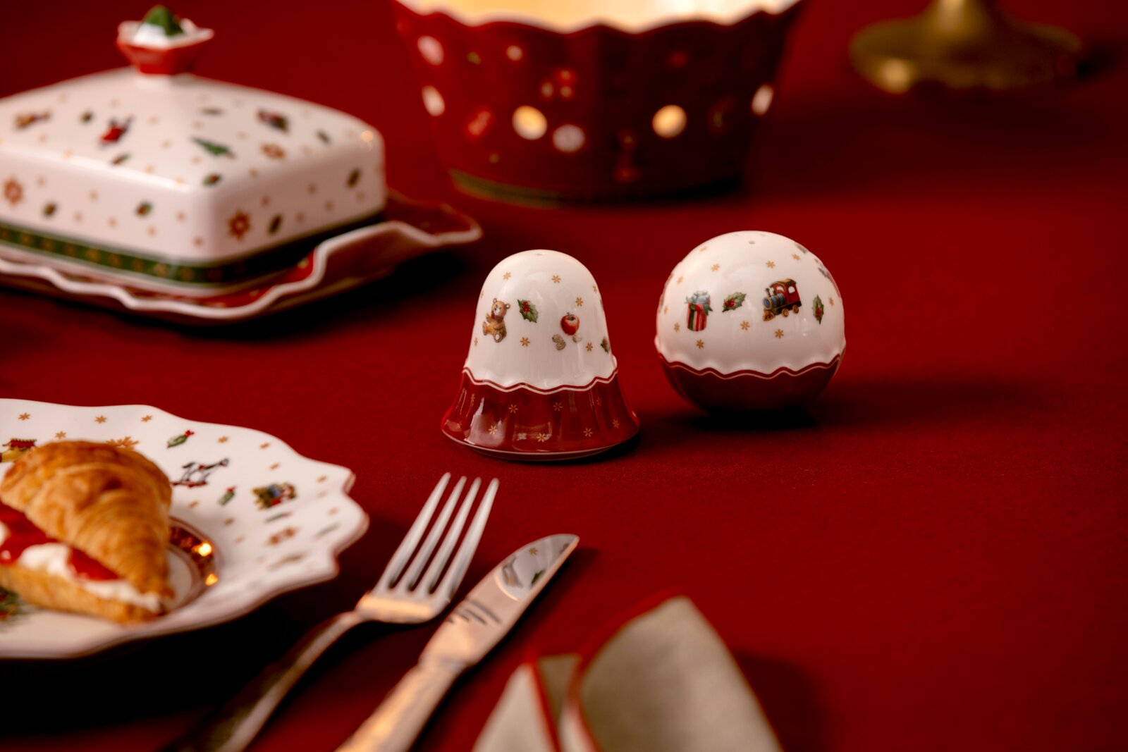 Weihnachtliches Salz- und Pfefferstreuer-Set aus weiß-rotem Porzellan mit Teddy-, Geschenk- und Spielzeugzugmotiven, in frontaler Dreiviertelansicht auf einer festlich gedeckten roten Tafel