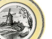 Dekorativer Porzellanteller mit gelbem Rand und schwarzem Zierdekor, in der Mitte schwarz-weiße Landschaftsszene mit Windmühle, Fluss und Boot, fotografiert in schräger Draufsicht (Detailansicht des rechten Tellerbereichs).