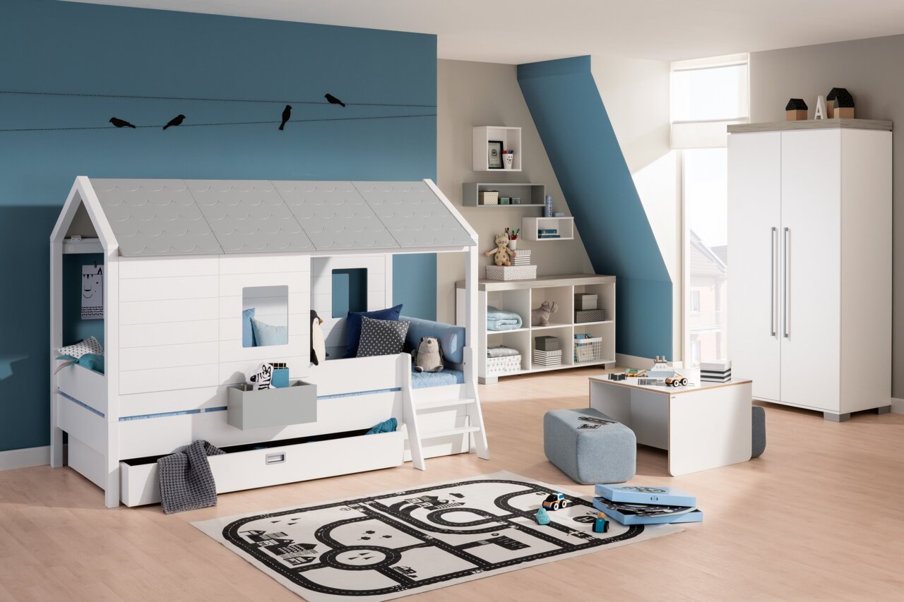 Paidi Dachschindel-Set TINY-HOUSE Kinderzimmer mit einem Bett in Form eines kleinen Hauses mit grauem Dach, umgeben von Regalen und Spielzeug, aus der Perspektive eines Raumes mit blauer Wand.