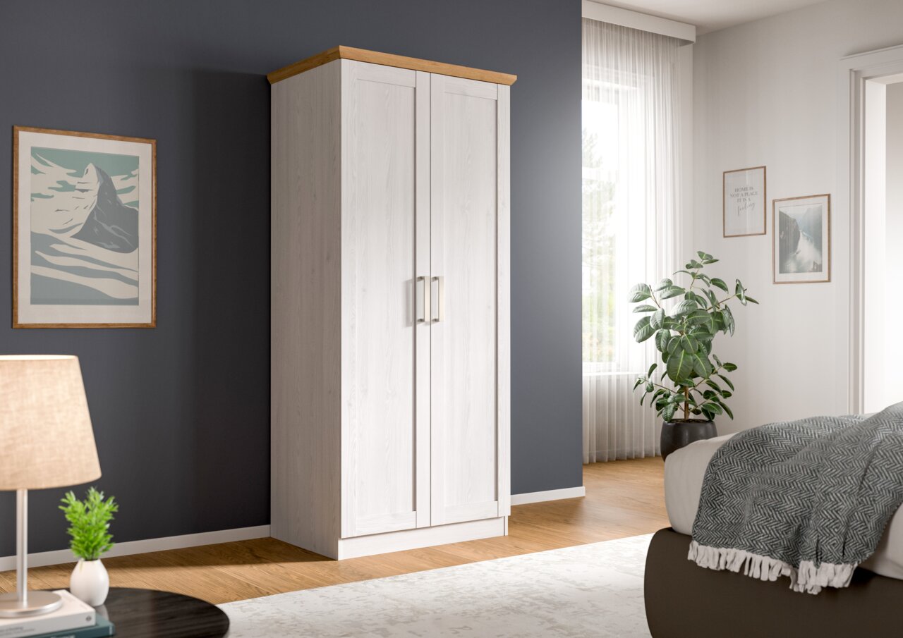 Zweitüriger Kleiderschrank in Weiß mit Holzkranz oben und silbernen Griffen, im Schlafzimmer vor dunkelgrauer Wand, perspektivisch in leichter Frontansicht von links.