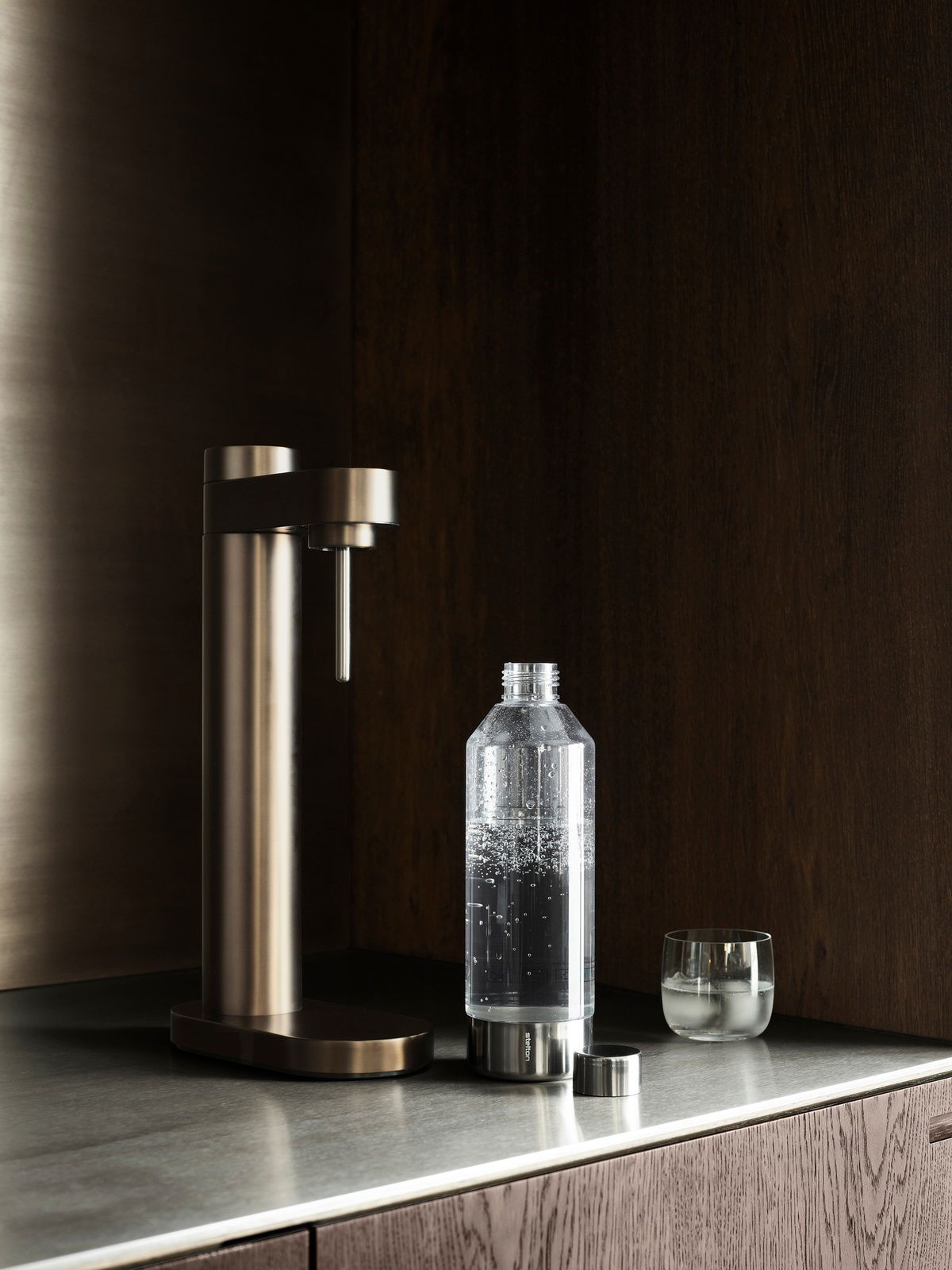 Stelton Wassersprudler BRUS in dunklem Braun auf einer Küchenarbeitsplatte, seitliche Perspektive. Daneben steht eine Flasche mit sprudelndem Wasser und ein Glas.