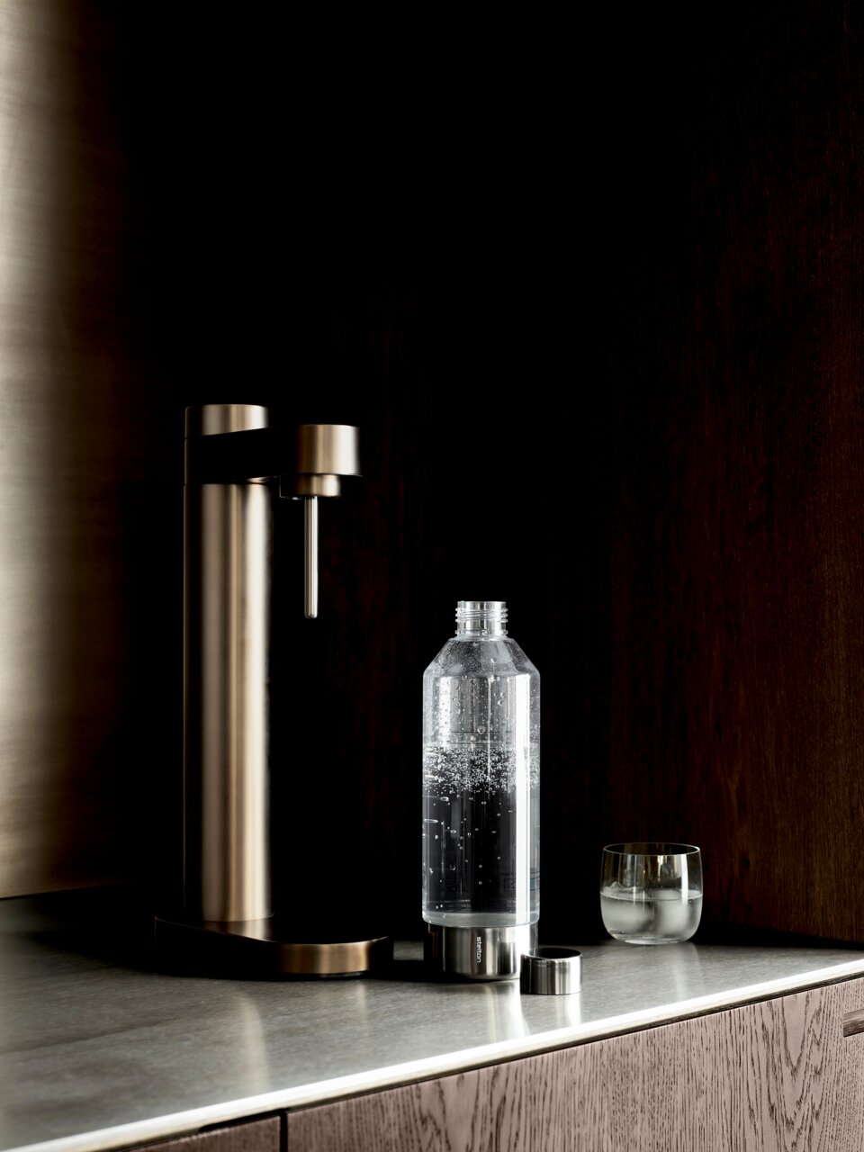 Stelton Wassersprudler BRUS Stelton Wassersprudler BRUS in dunklem Braun auf einer Küchenarbeitsplatte, seitliche Perspektive. Daneben steht eine Flasche mit sprudelndem Wasser und ein Glas.