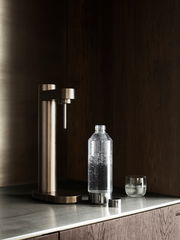 Stelton Wassersprudler BRUS in dunklem Braun auf einer Küchenarbeitsplatte, seitliche Perspektive. Daneben steht eine Flasche mit sprudelndem Wasser und ein Glas.