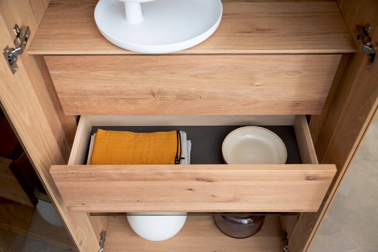 Geöffnete Schublade einer Vitrine aus Holz mit gelbem Tuch und weißem Teller, Ansicht von oben.