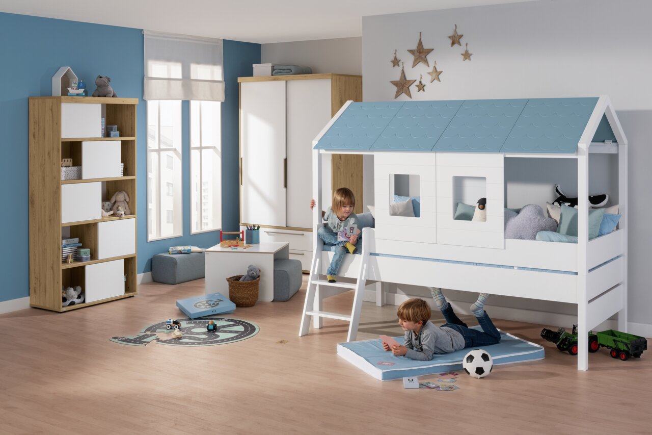 Paidi Dakleisteen TINY-HOUSE Kinderzimmer mit einem Hochbett in Form eines Hauses mit blauen Dachschindeln, zwei spielende Kinder, eines auf der Leiter und eines auf einer Matte darunter, Perspektive von vorne.