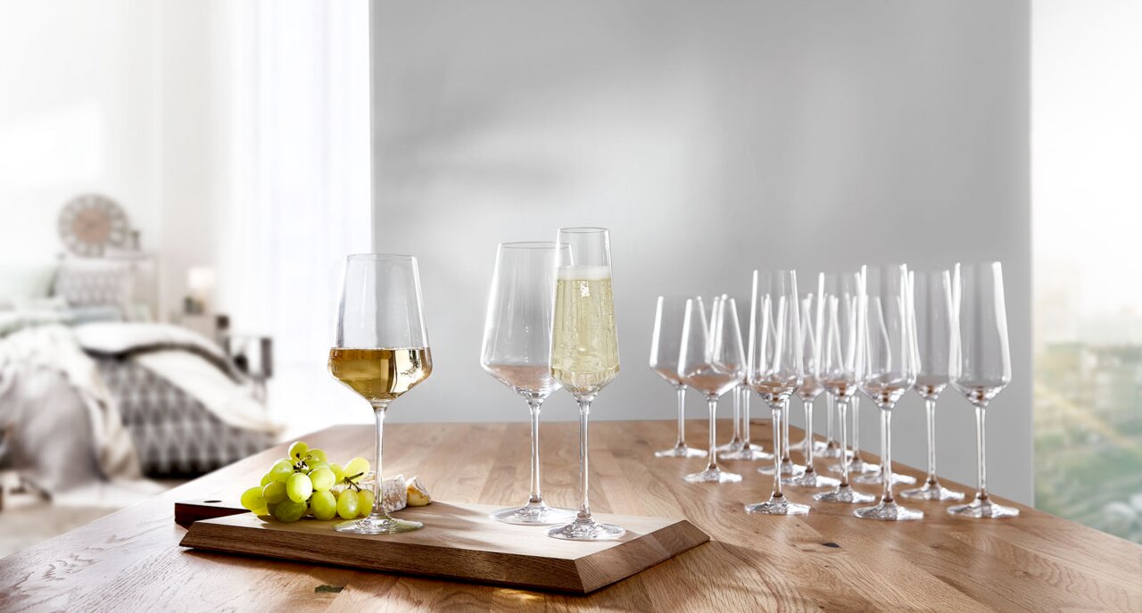 Kelchgläser-Set Puccini 18-tlg. auf einem Holztisch, mit gefüllten Gläsern im Vordergrund und einer modernen, hellen Raumkulisse im Hintergrund, seitliche Perspektive.