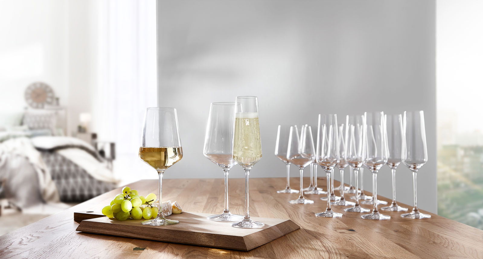 Kelchgläser-Set Puccini 18-tlg. auf einem Holztisch, mit gefüllten Gläsern im Vordergrund und einer modernen, hellen Raumkulisse im Hintergrund, seitliche Perspektive.
